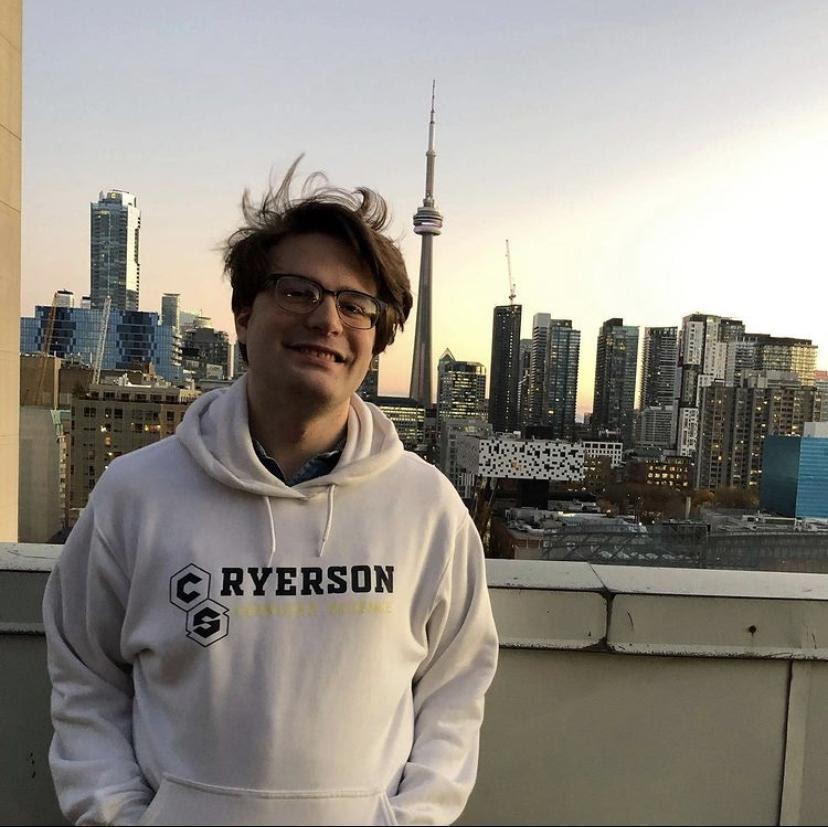 an individual smiles in front of a camera while standing on their balcony in the evening with the CN Tower in view behind them