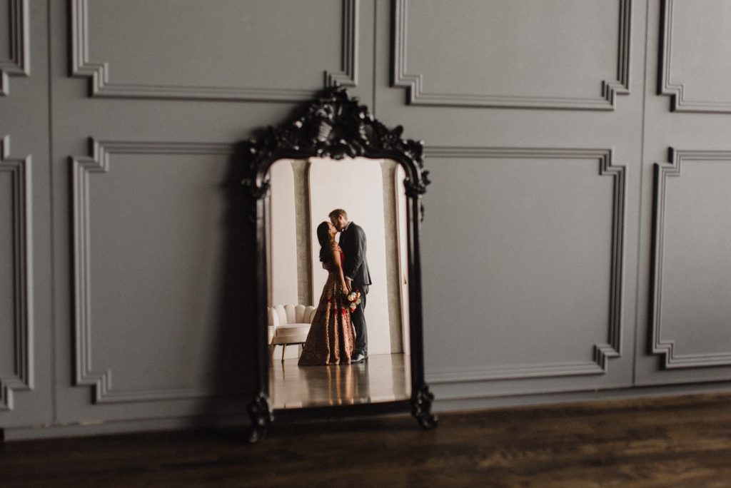 a mirror on a hardwood floor leaning on white walls with a full body shot of a couple looking at each other and holding each other closely visible in the mirror. the woman is wearing a traditional South Asian red dress while the man is wearing a suit and tie