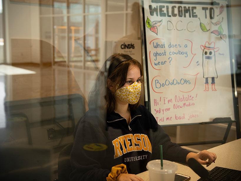 a girl in a Ryerson hoodie sits at a desk behind a plexi glass wearing a mask