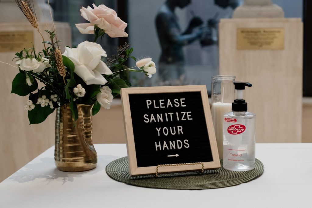 a board that says "Please sanitize your hands" on a table used at weddings with flowers and hand sanitizer surrounding the board