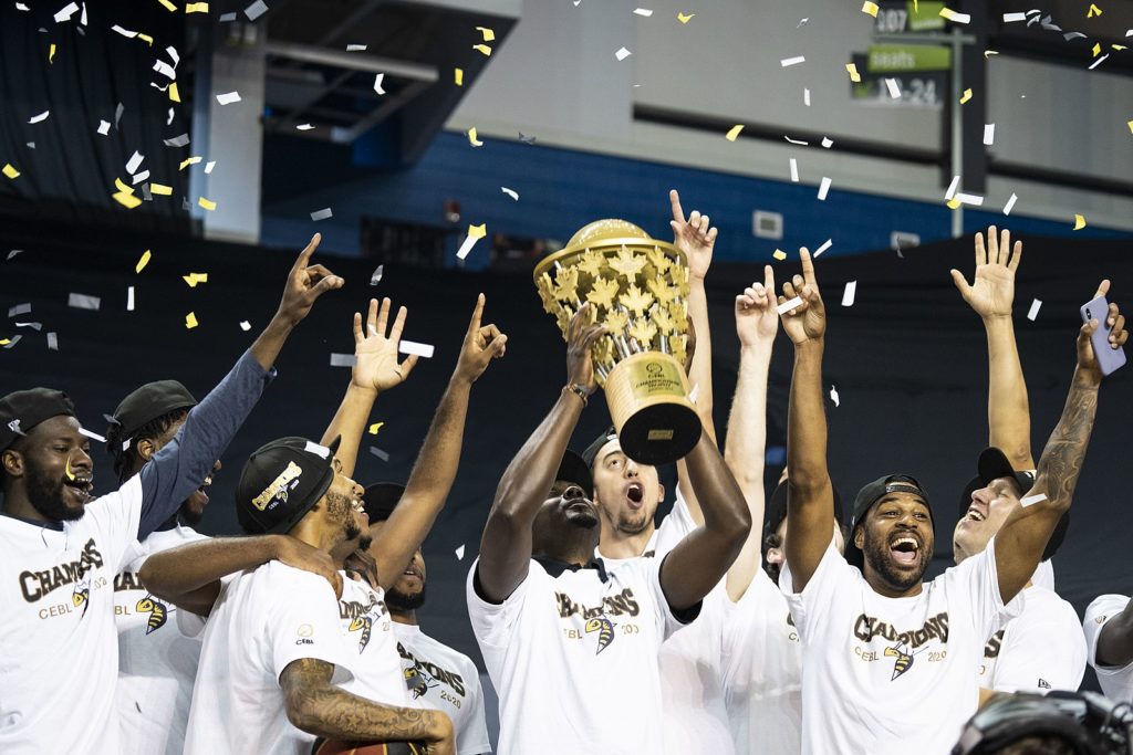 A group of basketball players celebrate a championship win, holding a trophy with confetti falling.