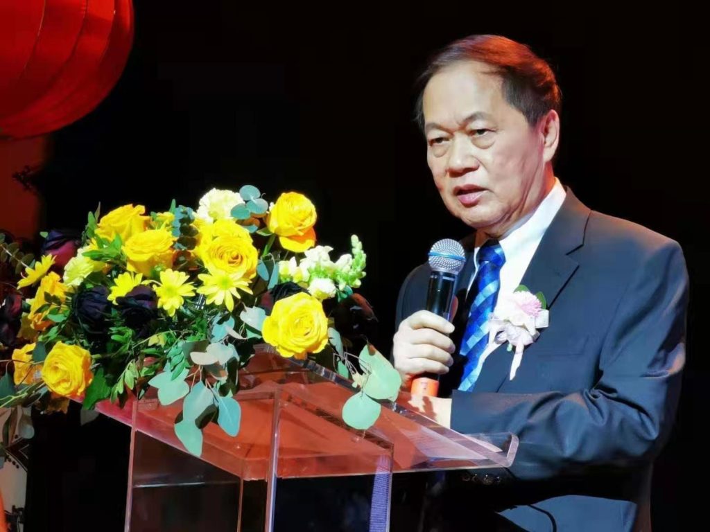 an elderly man dressed in a blue suit and tie stands in front of a podium with yellow flowers and speaks into a mic