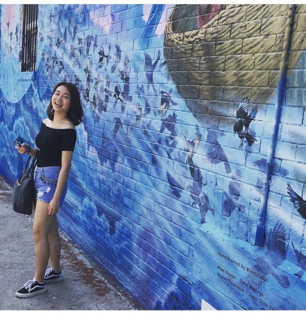 a girl stands in front of a blue brick wall outside and smiles at the camera while holding a purse