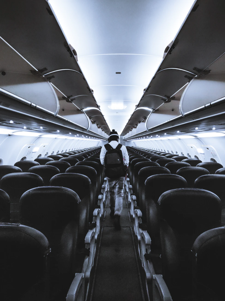 a view from the middle of an airplane aisle with rows of black and gray seats on the left and right sides and a traveller with a backpack walking away from the camera