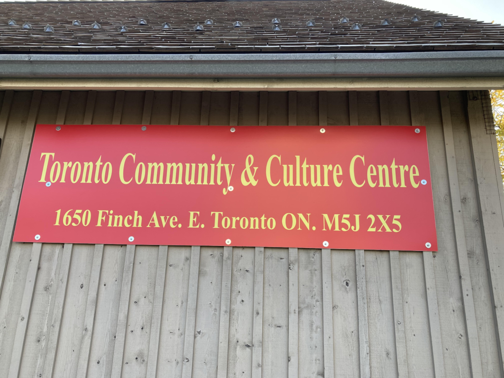 A red sign on a wooden wall with the phrase "Toronto Community and Culture Centre at 1650 Finch Avenue East" written in gold