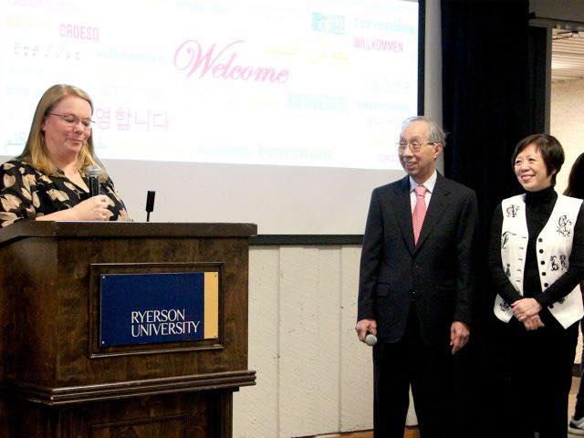 a woman speaks at a podium into a mic with two people on the right side watching her as she speaks