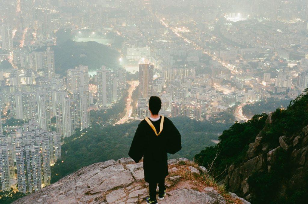 a boy wearing a cap and gown stands on a cliff looking out at the city