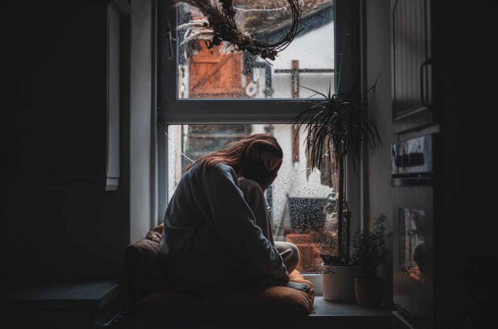 A photo of a person wearing a mask, sitting at a window seat holding their head.