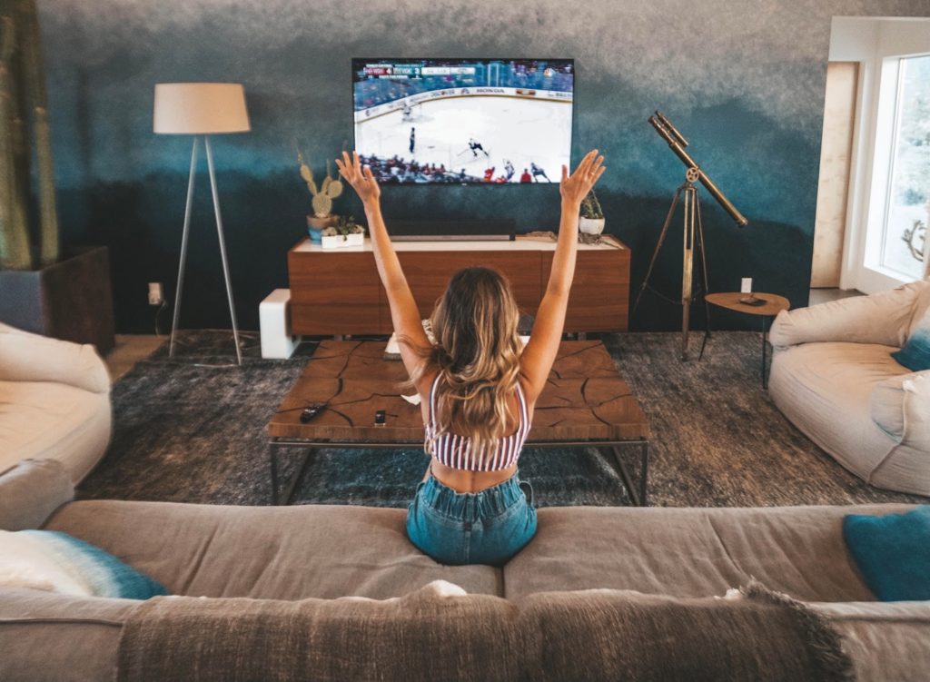 a woman puts her hands up as she sits on a couch looking at a tv screen with a hockey game on it