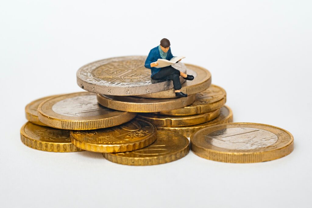 A photo/illustration of a small model of a human holding an open newspaper, positioned on a pile of coins.
