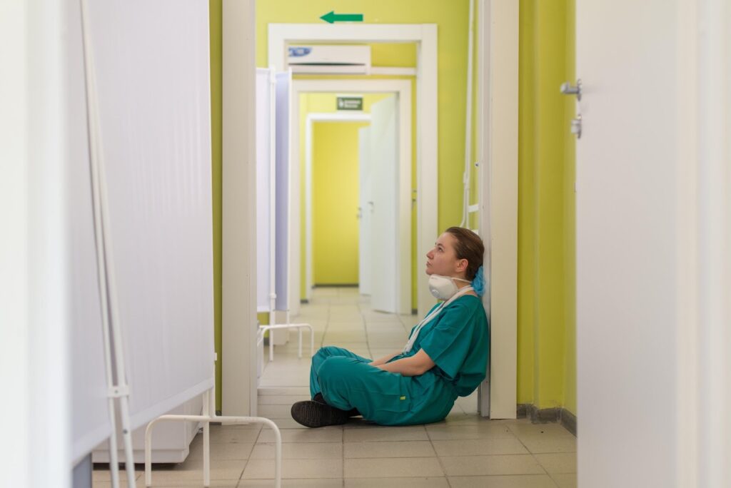 Photo of a person dressed in medical garb/scrubs wearing a mask lowered to their chin, sitting in a lime green hallway that could be a hospital.