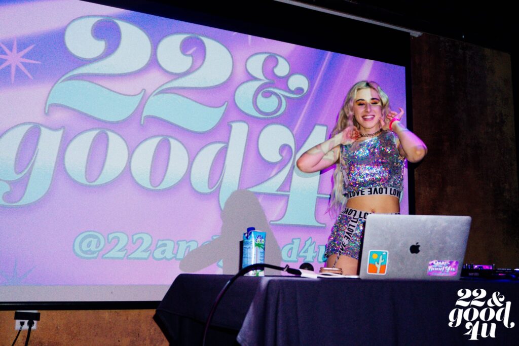 A DJ wears a sparkling sequinned outfit and stands next to a projector that reads "22 and good 4 u"