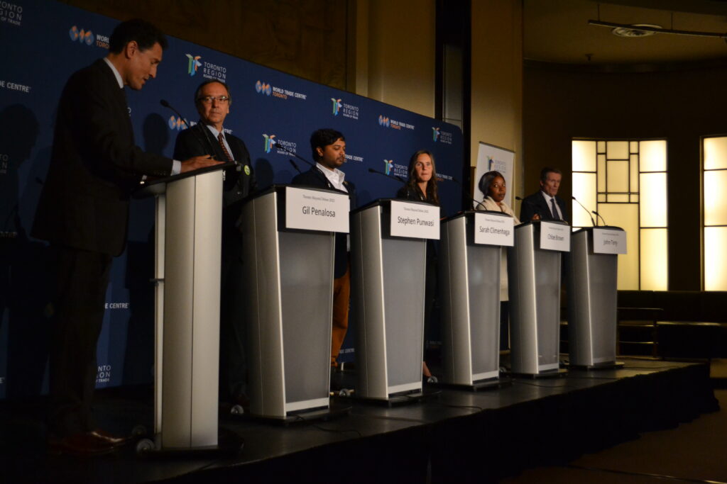 Steve Paikin and the five mayoral candidates standing at podiums side by side, at the Toronto mayoral debate on Oct. 17, 2022.