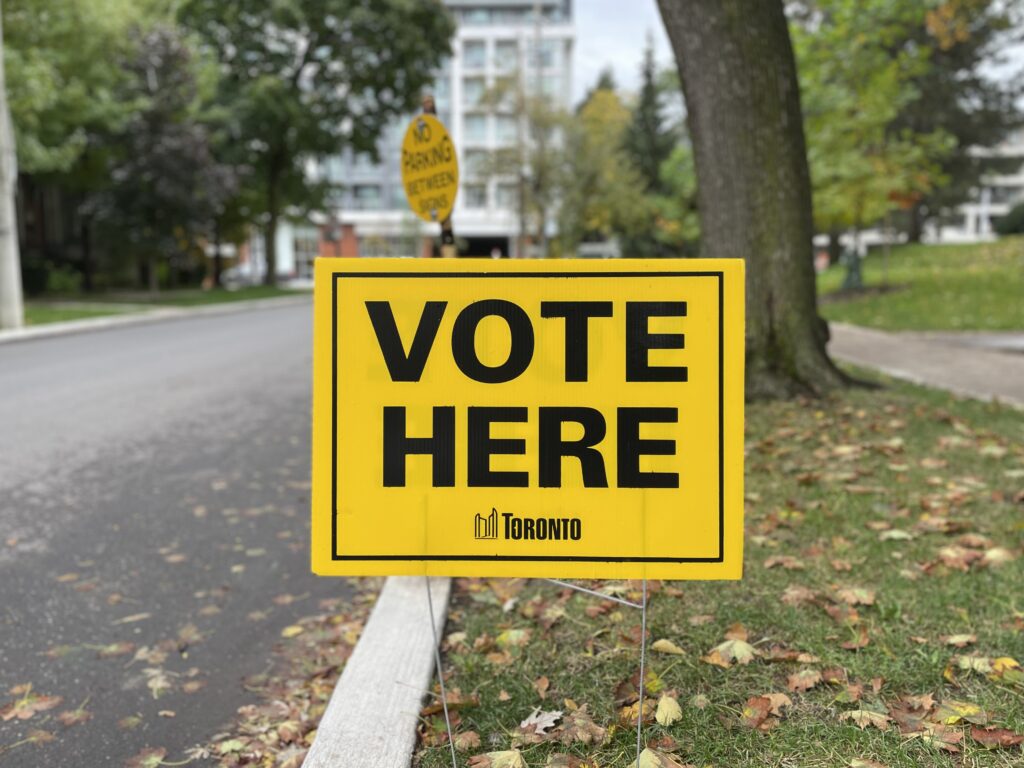 A "VOTE HERE" sign from the City of Toronto is pictured on a lawn in fall.