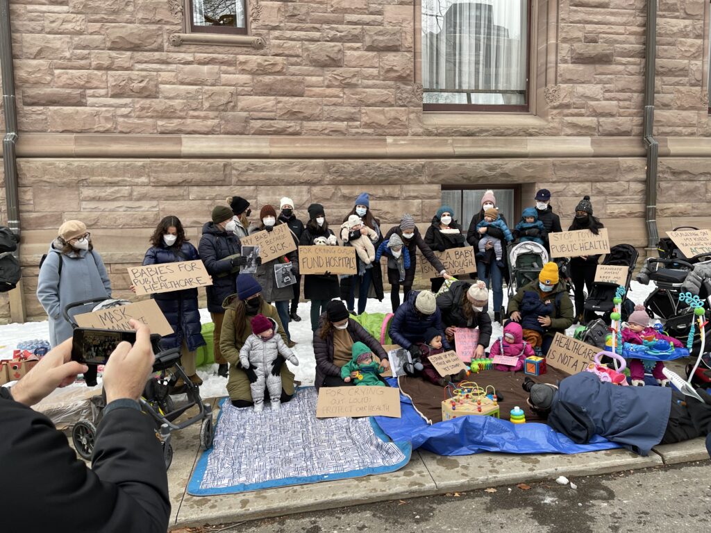Image of a large group of parent protestors with their children holding signs outside of Toronto's Ontario Legislative Building.