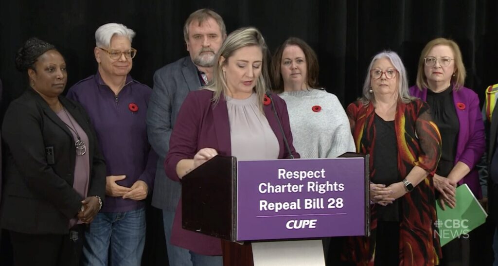 Ontario School Board Council of Unions president Laura Walton speaks at a press conference, lined up behind her are various private and public sector union representatives.