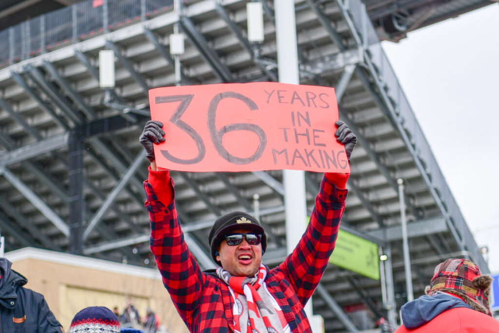 Fan holding a sign with writing. Wearing black sunglasses, tuque, and plaid shirt. 