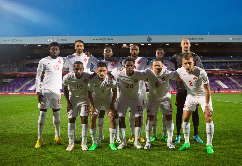 Canadian Men's soccer team in white kits on turf field. 