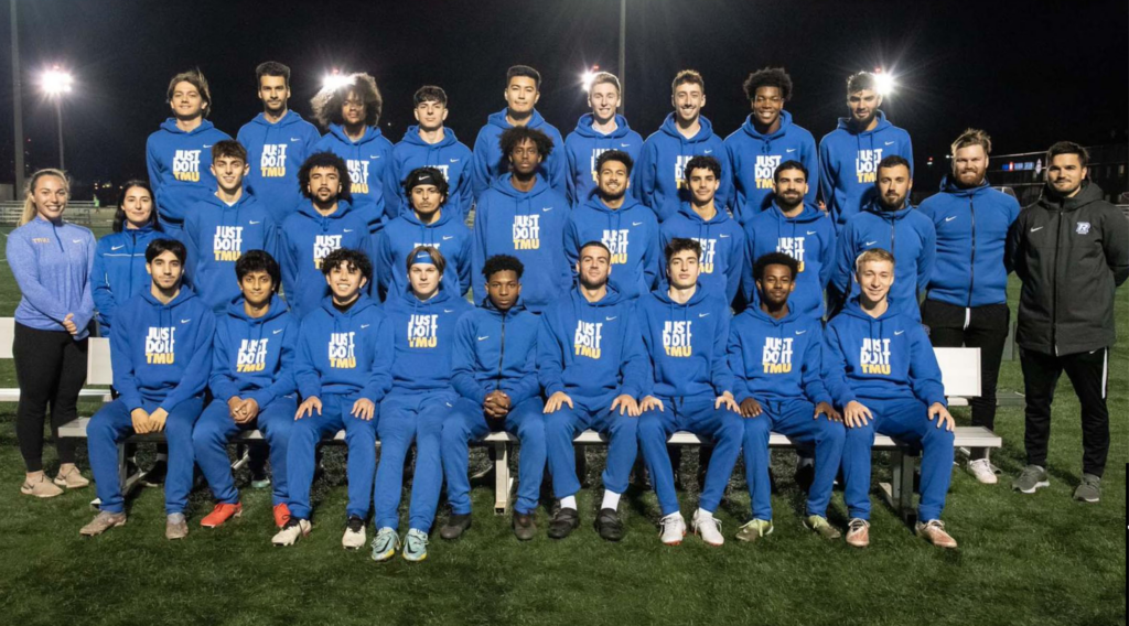 TMU Men's soccer team sitting at a bench for squad photo in the park.