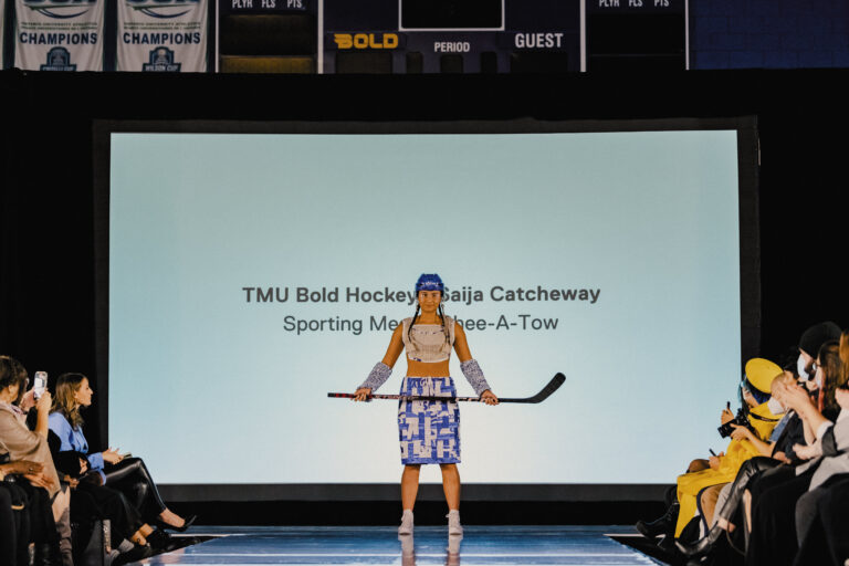 a TMU student-athlete models an upcycled outfit made of old Ryerson-branded materials with a hockey stick on a black stage