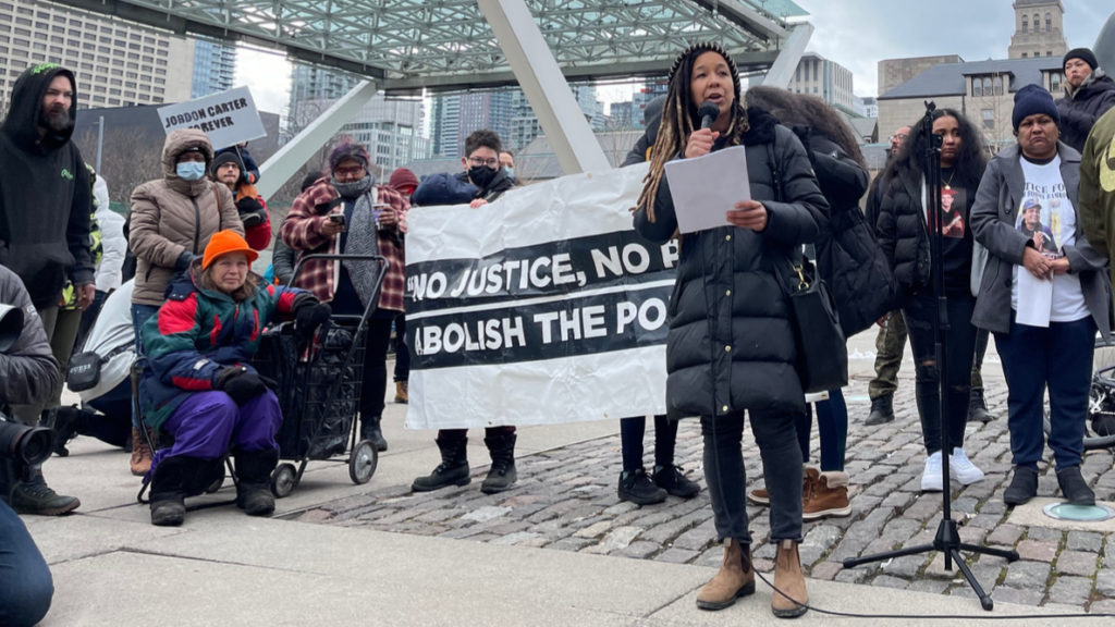 A woman in winter clothes holds a microphone in one hand and a sheet of paper in the other. She speaks into the microphone, addressing the crowd of people in front of her.