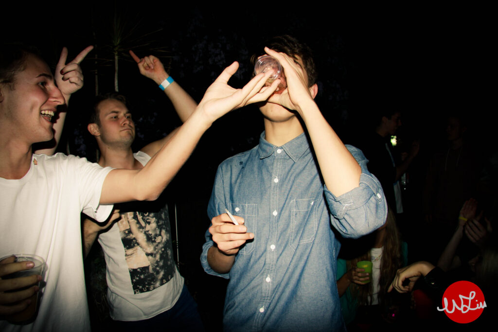 A student in a white t-shirt pushes the bottom of another student's drinking glass up, forcing him to drink faster. The student drinking is wearing a blue denim shirt and holds a cigarette in his other hand. Another student in a white t-shirt stands behind the other two, dancing with his hands in the air.