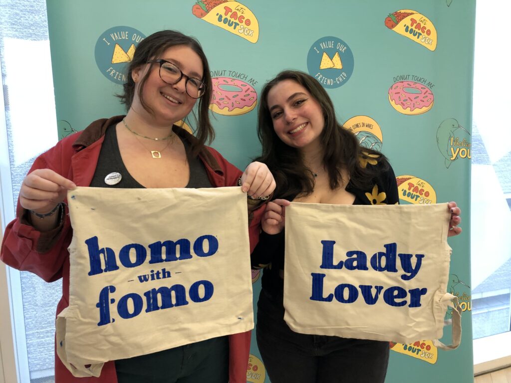 Two students stand side by side behind a patterned screen that has a turquoise background. They are holding tote bags with fresh blue lettering on them. The tote bag on the left reads "homo with fomo", and the tote bag on the right reads "Lady Lover".