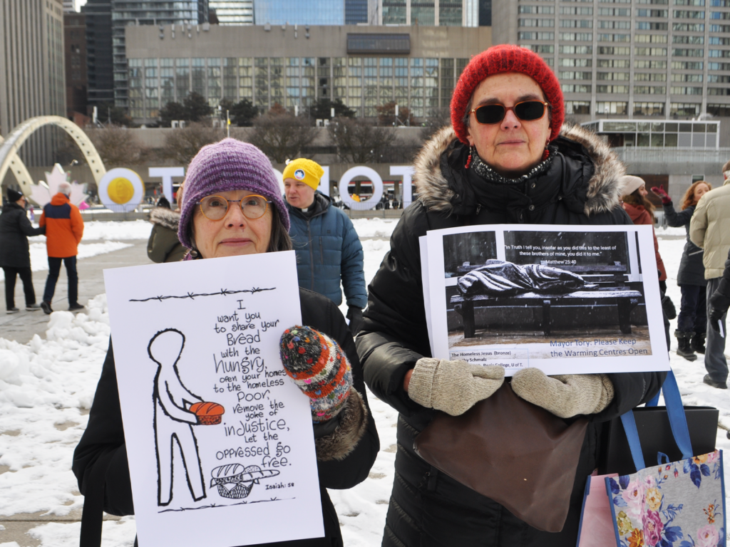 Two individuals stand in Nathan Phillips Square in Toronto Ontario both holding signs. The left sign says "I want you to share your bread with the hungry, open your homes to the homeless poor, remove the yoke of injustice, let the oppressed go free. Bible quote from Isaiah: 58," with an image of a illustrated person holding a loaf of bread. The right poster is a black and white photo of a body silhouette wrapped in a blanket laying on a bench with the text "In Truth I tell you, insofar as you did this to the the least of these brothers of mine, you did this to me. Matthew 25:40" as well as "Mayor Tory: Please Keep The Warming Centres Open."