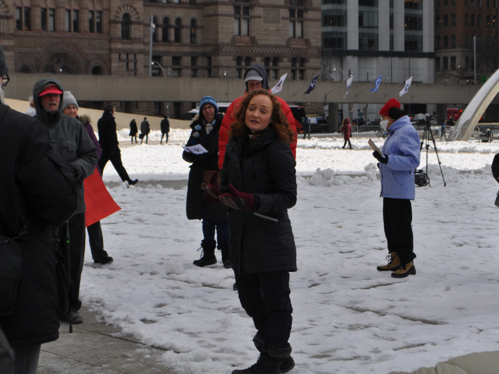 Alexa Gilmore stands in the centre of the photo taken at a snowy Nathan Phillips Square in Toronto. She is speaking to people standing around and listening to her. 