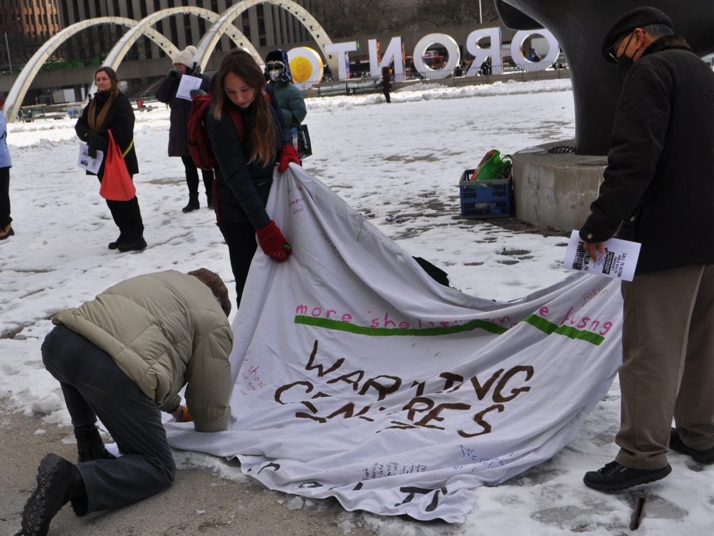 An individual is bent over on one knee writing on the large cloth sign that says "Warming Centres 24-7." The cloth is being held up by two people looking at what the individual is writing. 