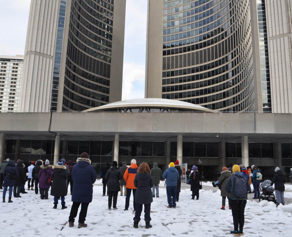 Taken from behind, the photograph shows around a dozen people bundled up in winter jackets facing Toronto City Hall. 