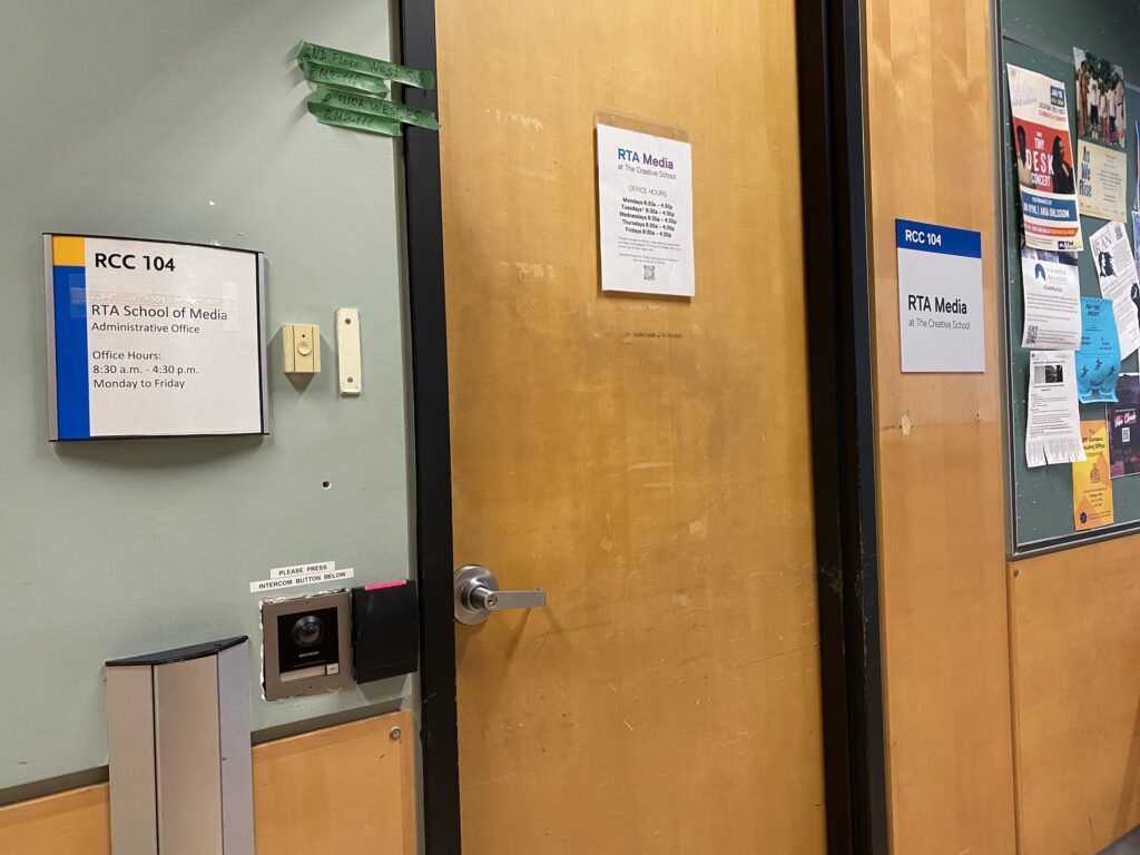 A sign next to a door that reads: RCC 104 RTA School of Media Administrative Office. Office Hours: 9:30 a.m. to 4:30 p.m. Monday to Friday. Below is a camera intercom system next to a key card reader. On the right of the door there is a sign that reads: RCC 104. RTA Media at The Creative School. 