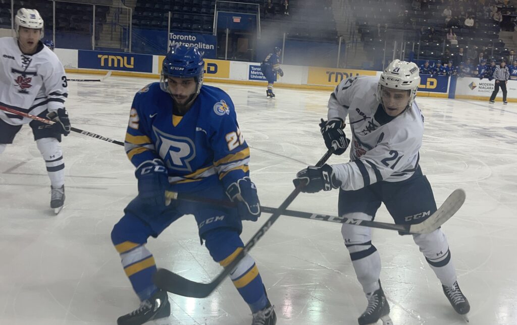 A TMU Bold Hockey player and a U of T Hockey player look at boards, with their sticks deadlocked