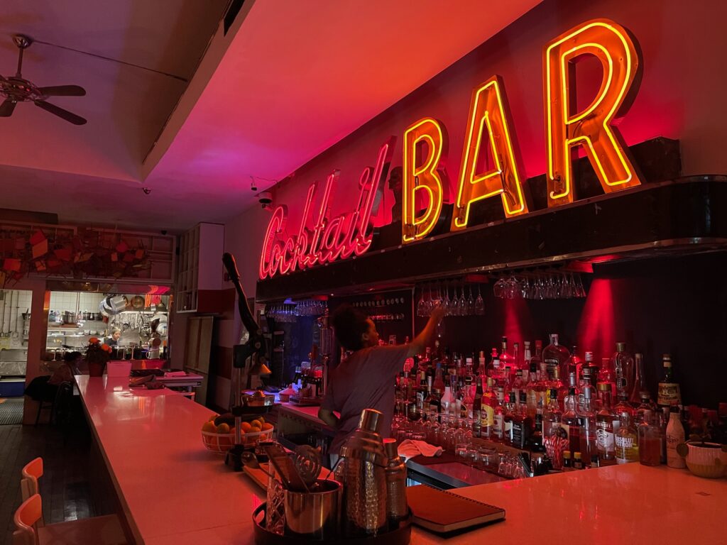 A neon sign above the bar area in a restaurant reads "cocktail bar." Cocktail is in red letters and bar is lit up in yellow. An employee in a grey t-shirt puts away wine glasses in the bar.