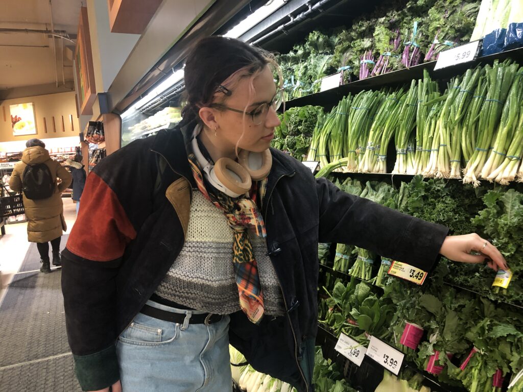 Sofia Stadler reaches towards a shelf of leafy greens while two shoppers can be seen walking behind them. 