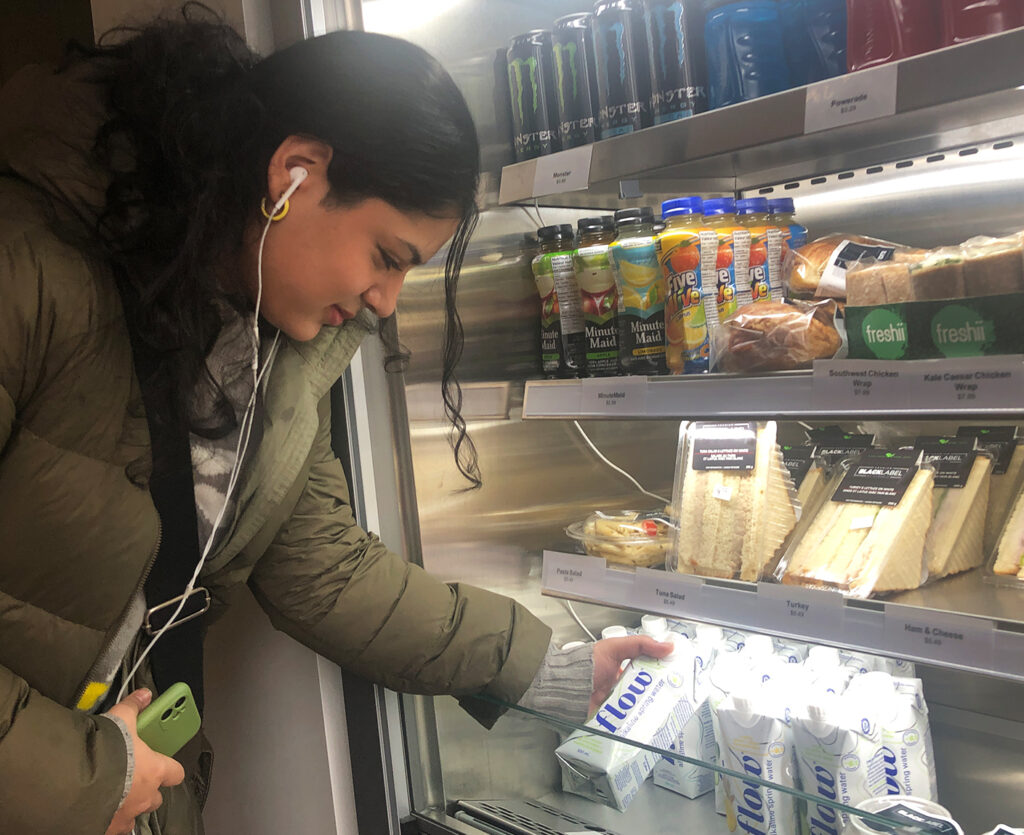 Zahra Ali stands to the left of a refrigerated merchandiser and picks out a drink.