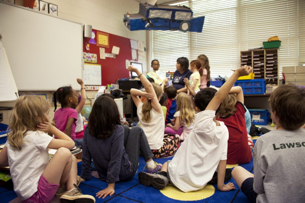 A group of children sit on the floor facing a teacher, a few with raised hands, in a classroom setting.
