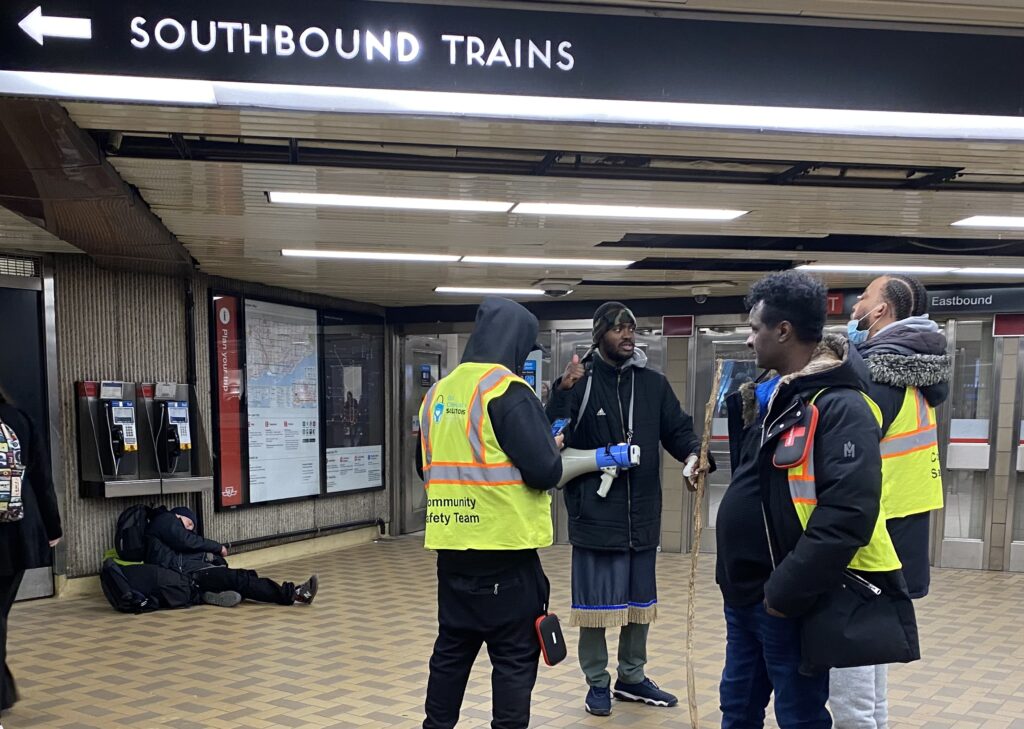 Three community safety ambassadors stand in a group with neon jackets on. An unhoused man sleeps on the floor near them.