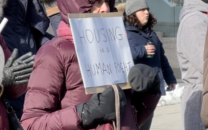 A woman in a red winter coat holds up a white sign with black writing that reads "Housing is a human right."