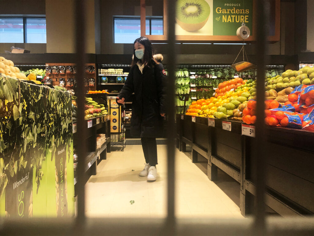 A shopper wearing a black long winter coat and a medical mask walks through an aisle with fresh produce on either side. 