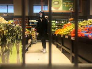 A shopper wearing a black long winter coat and a medical mask walks through an aisle with fresh produce on either side.
