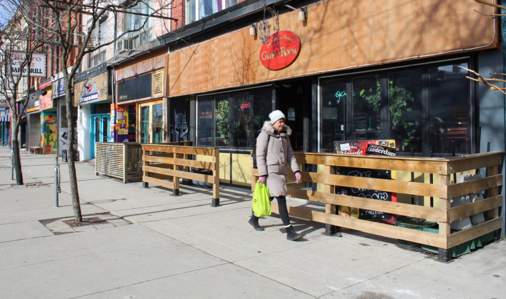 A woman in a winter jacket holds a green grocery bag and walks past a row of restaurants.