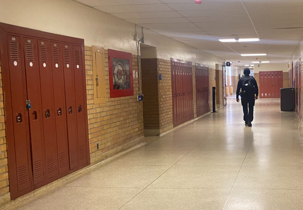 A security guard with his back turned to the camera walking down a hallway with lockers on either side.
