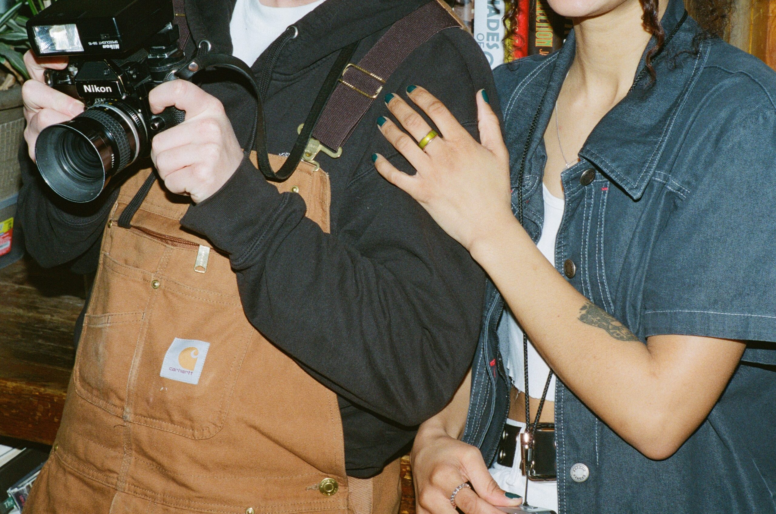 Two people stand in front of the camera in brown Carhartt overalls and a short sleeve denim shirt. One of them takes a photo with a Nikon camera. 