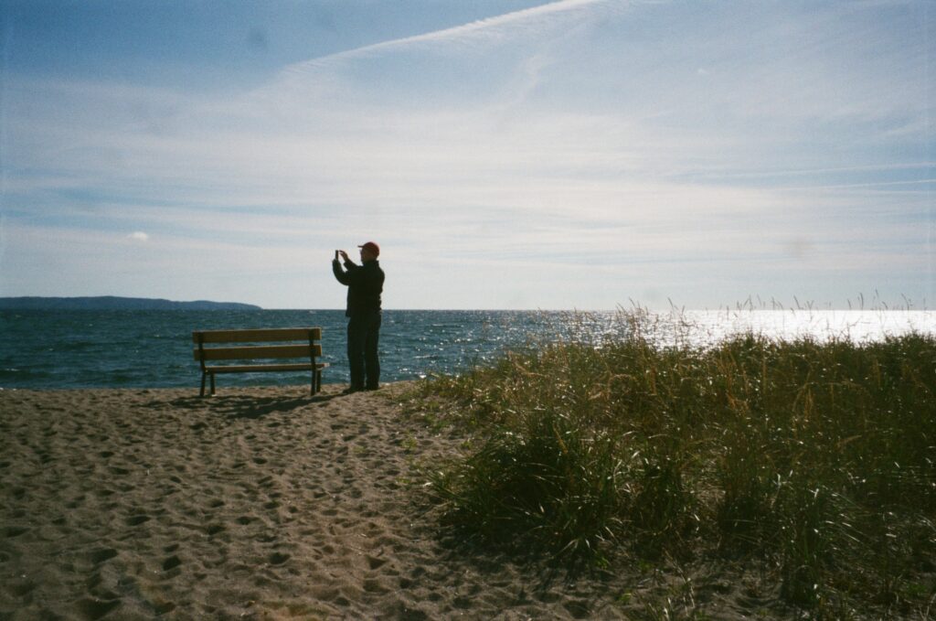 A man stands in front of water on a beach to take a photo. 