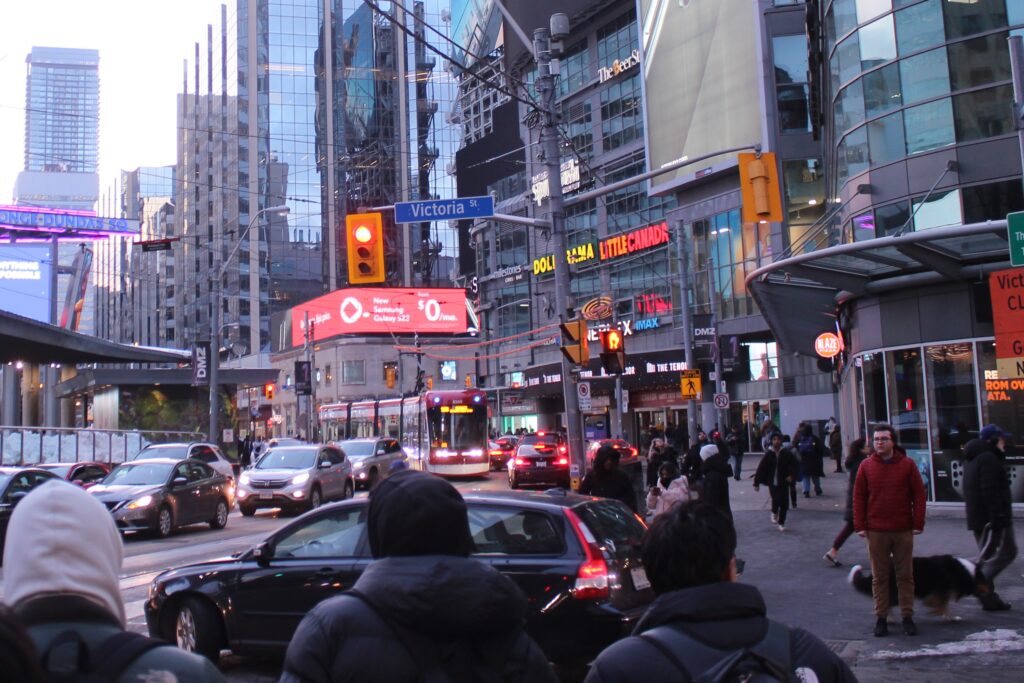 Three people face an intersection near Dundas Square. Cars and a streetcar are on the street, with bright lights from buildings above.