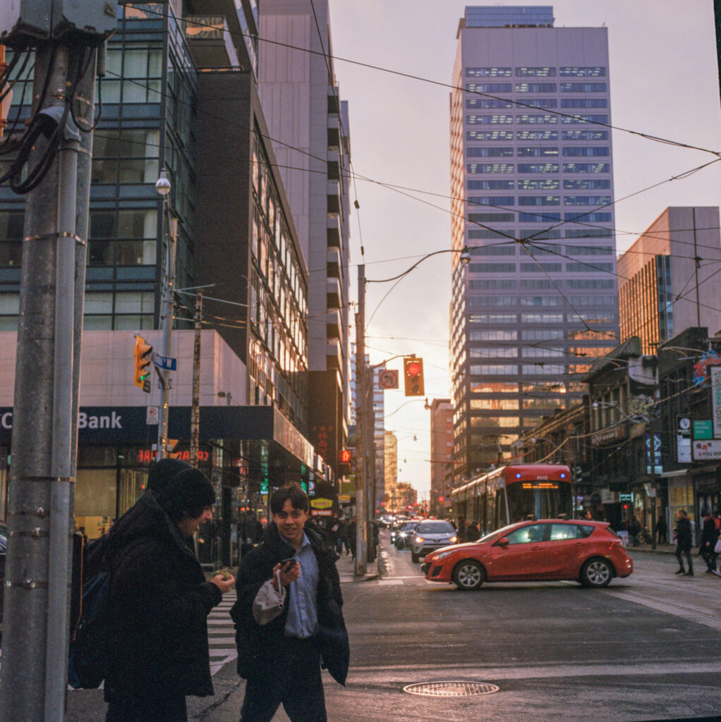 Three people prepare to cross the road inToronto as the sun sets. A red car drives past the street with apartment buildings in the background.