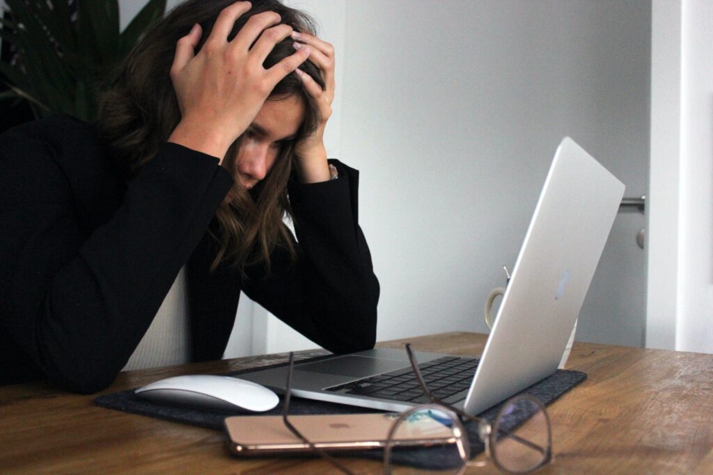 A young person hands their head in their hands while sitting at a desk in front of a laptop. 