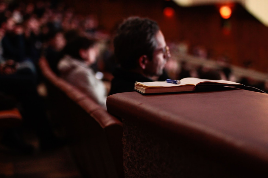 Photo focused in on a book with a pen with an audience of people in the background