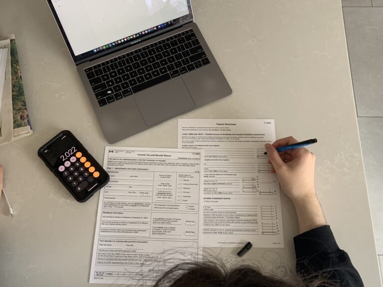 A student holds a pen while looking at an income tax form, a calculator, and a computer.